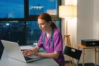 A women in scrubs sits at her desk while typing on a her laptop